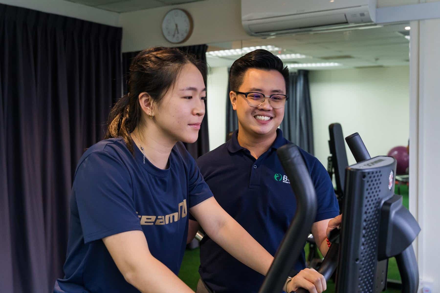 Physiotherapists discussing patient progress near stationary bike in exercise area. Shows cardio rehabilitation, equipment-based therapy, monitoring, and exercise supervision in clinic facility. Keywords: cardio equipment, stationary bike, exercise supervision, rehabilitation program, patient monitoring, fitness facility.