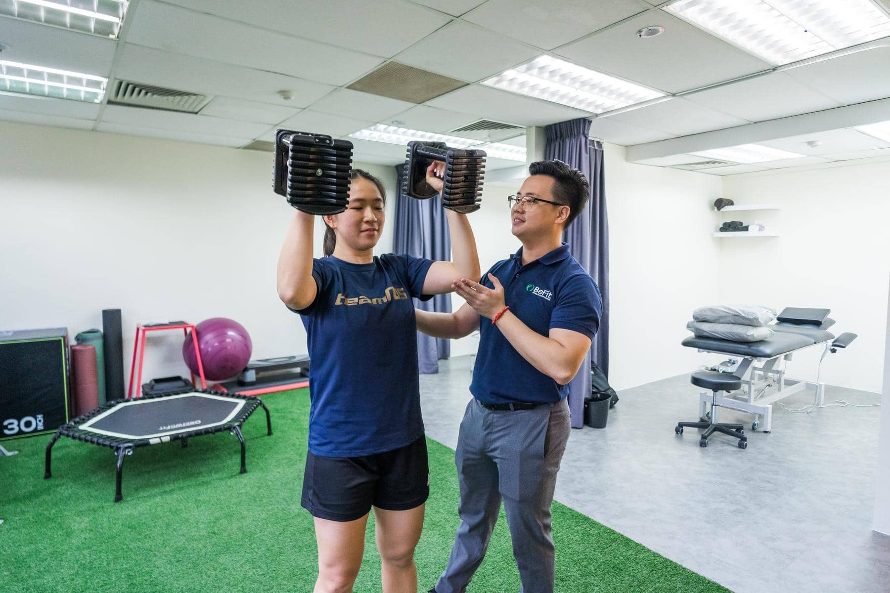Patient performing dumbbell weight lifting exercise with therapist guidance and supervision in clinic gym. Demonstrates strength conditioning, resistance training, progressive rehabilitation, and exercise coaching. Keywords: strength training, weight lifting, exercise guidance, resistance program, rehabilitation exercise, supervised training.